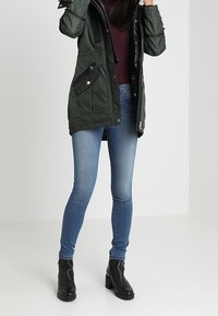 Parka verde imbottita con cappuccio, cerniera e multiple tasche. Abbinata a jeans blu aderenti e stivaletti neri alla caviglia. Texture liscia.