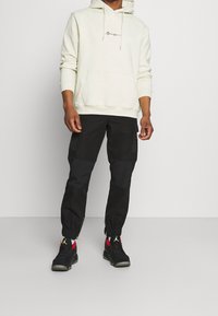Hoodie beige chiaro con tasca frontale e cordini, abbinato a pantaloni cargo neri con polsini con zip e tasche laterali, completato da scarpe nere.