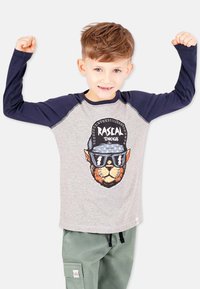 Grå langærmet skjorte med marineblå ærmer, prydet med et grafisk motiv af en løve iført solbriller og en kasket. Teksten nedenfor lyder "Rascal Tendencies."