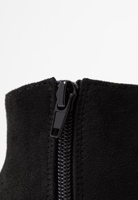 Gros plan sur une botte en daim noir, dotée d'une fermeture éclair noire brillante longeant verticalement le côté, sur un fond clair et uni.