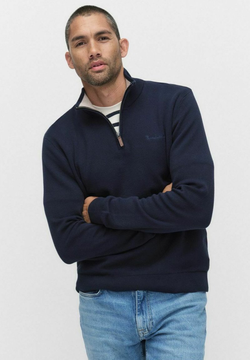 Marinblå zip-up sweatshirt med ribbade muddar och nederkant. Har en krämfärgad inre krage och bärs över en randig crewneck-tröja.