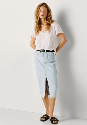 Femeie cu păr lung și creț purtând un tricou alb cu decolteu în V, fustă midi din denim albastru deschis cu slit frontal, curea neagră și sandale negre cu ținte.