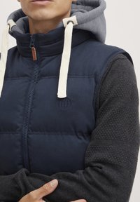 Navy blue padded vest with a high collar, gray hood, cream drawstrings, and textured black sleeves featuring a circular logo on the chest.