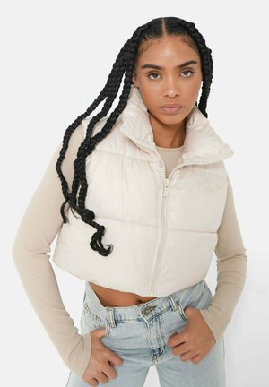 Lichtbeige, kort puffer vest met hoge kraag, rits aan de voorkant en geborduurde secties. Gecombineerd met een ribbelige beige langemouwtop en blauwe jeans.