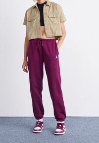 Geknipte beige shirt met twee borstzakken, gecombineerd met losse paarse sweatpants met een wit logo en elastische boorden, gedragen met bordeauxrode sneakers.