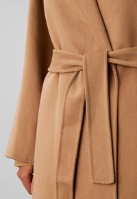 Gros plan d'une personne portant un manteau en laine couleur camel, noué à la taille avec une ceinture assortie, avec une main partiellement visible.