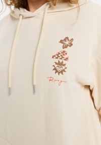 Beige hoodie met trekkoord; voorzien van bruine bloem- en patroon grafieken en een rode "Roxy" logo op de voorkant. Gladde stoftextuur.
