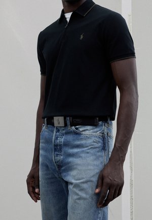 Homme portant un polo noir rentré dans un jean bleu clair avec une ceinture noire, debout devant un mur clair uni.