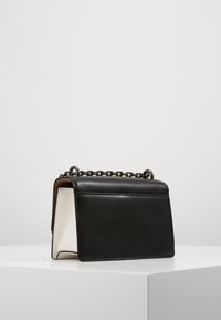 Bolso de mano de cuero negro con interior blanco, que cuenta con un asa de cadena y una forma estructurada. Cierre de solapa superior con costuras decorativas.