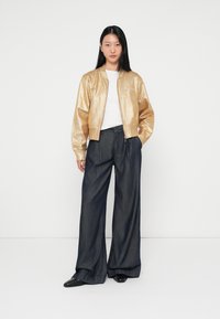 Jachetă bomber metalizată aurie, cu închidere cu fermoar, peste o cămașă albă, asociată cu pantaloni cu picioare largi din jeans închis la culoare. Pantofi negri finalizează ținuta.