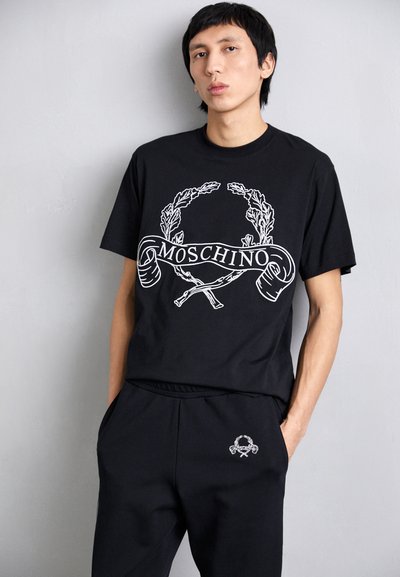 Must have musta t-särk, millel on valge graafiline disain laurioksi ja "Moschino" tekst, koos sobivate mustade dressipükstega.