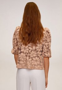 Vaaleanpunainen pitsipusero, jossa on puhvimaiset lyhyet hihat, kukkakuviointi ja lyhyt, vartalonmyötäinen leikkaus, valmistettu yksityiskohtaisesta pitsimateriaalista.