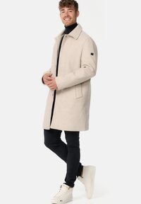 Lys beige uldfrakke med krave, sidelommer og en sort detalje på ærmet, parret med mørke bukser og hvide sneakers.