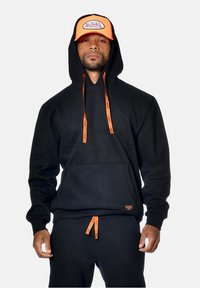 Haut à capuche noir avec des cordons de serrage orange et des détails de poche assortis. Associe à un pantalon noir, porté avec une casquette à visière orange arborant un logo.