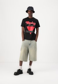 Camiseta negra con gráfico rojo de "Replay", pantalones cortos de mezclilla de lavado claro, zapatos negros gruesos y un sombrero estilo bucket oscuro, de pie frente a un fondo blanco.
