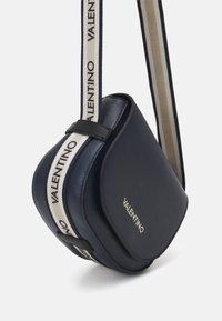 Granatowa torebka na pasku o eleganckim, zaokrąglonym kształcie, wykonana z gładkiej skóry. Posiada złote logo oraz regulowany pasek w wzory.