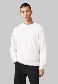 Felpa bianca con girocollo, polsini e orlo a coste, con un piccolo logo in basso a sinistra. Il materiale sembra morbido e textured.