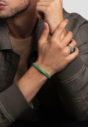 Green woven bracelet made of beads, wrapped around a wrist. Notable silver accents and a contrasting textured outfit.