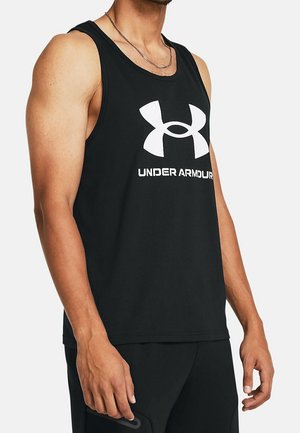 Mężczyzna w czarnym, bezrękawnym podkoszulku Under Armour i czarnych szortach, stoi z rękami luźno opuszczonymi wzdłuż ciała na białym tle.