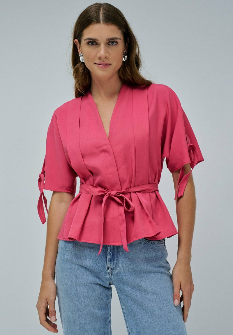 Top rosa con mangas cortas, detalles de lazo, escote en V y volante. Combinado con jeans azul claro para un look casual.