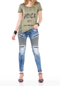 Camiseta verde de manga corta con la impresión "TÚ ROCKEAS MI CIPO Y BAK"; jeans de mezclilla azul con un patrón a rayas y detalles con cremalleras; sandalias de tacón alto.