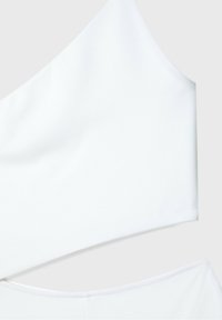 Crop top asymétrique blanc avec une bretelle large sur fond neutre.