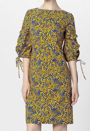 Persoon met een knielange jurk met geel en blauw bladmuster, driekwart geplooide mouwen en gestrikte strikjes aan de ellebogen.