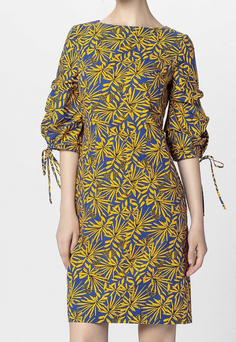Personne portant une robe longueur genou à motif de feuilles jaunes et bleues, manches trois-quarts froncées et nœuds attachés aux coudes.
