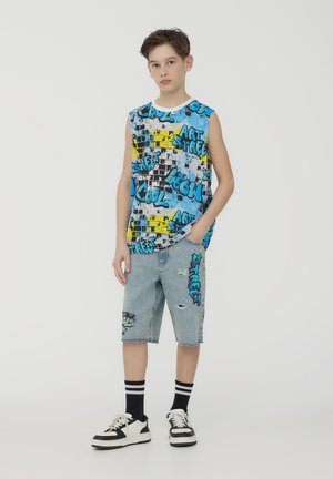 Canotta blu con grafiche caratterizzata da motivi a schizzo neri e gialli; abbinata a pantaloni denim strappati e sneakers nere e bianche.