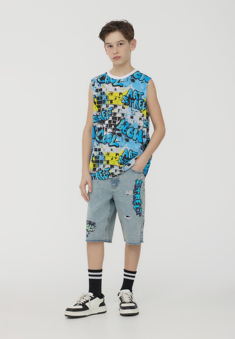 Canotta blu con grafiche caratterizzata da motivi a schizzo neri e gialli; abbinata a pantaloni denim strappati e sneakers nere e bianche.