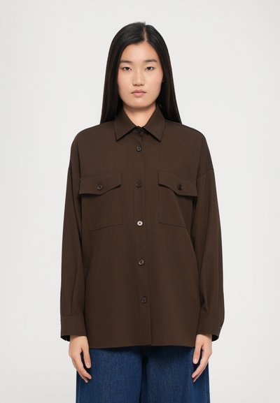 Chemise brun à boutons avec deux poches poitrine, dotée d'un col et de manches longues, en tissu lisse, associée à un pantalon en denim bleu.