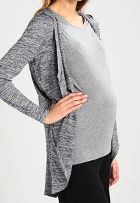 Šedý cardigan s dlouhým rukávem a kapucí, který má melírovanou texturu, vrstvený otevřený předek, nošený přes šedý top a černé kalhoty.