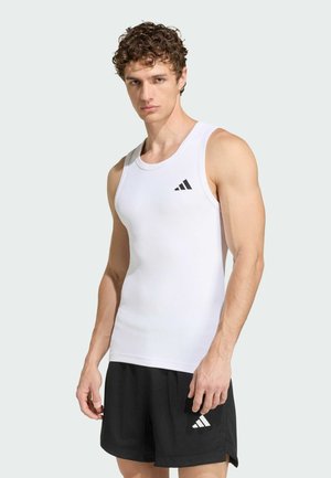 Giovane uomo che indossa una canottiera bianca senza maniche Adidas e pantaloncini neri Adidas, in piedi davanti a uno sfondo chiaro e semplice.