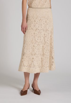 Beżowa koronkowa spódnica midi w kwiaty noszona z brązowymi butami na niskim obcasie o spiczastym nosku, pokazana od talii do stóp na jednolitym tle.