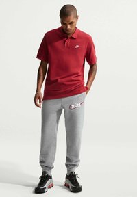 Bărbat purtând un tricou polo roșu Nike, pantaloni de trening gri Nike și adidași Nike în alb și negru, stând cu o mână în buzunar.