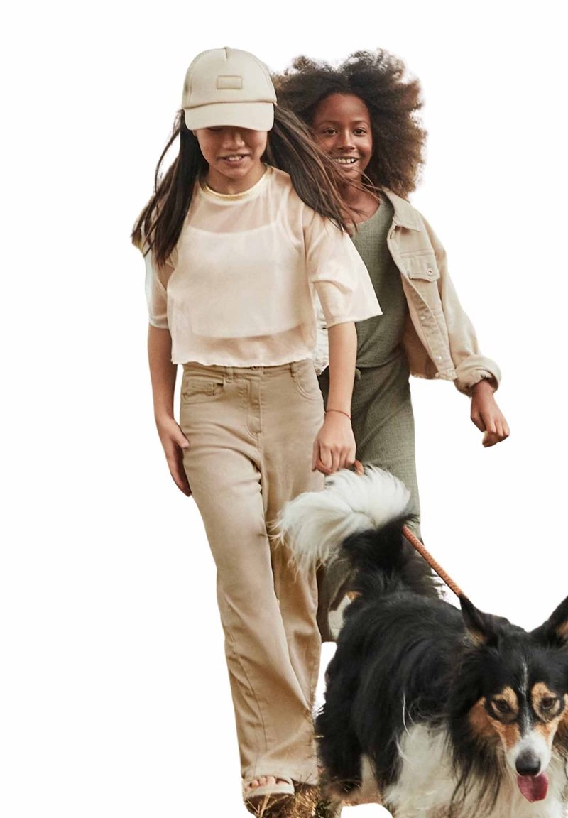 Due bambini camminano con un cane bianco e nero. Uno indossa un top trasparente beige e pantaloni, mentre l'altro sfoggia una tuta verde e una giacca.