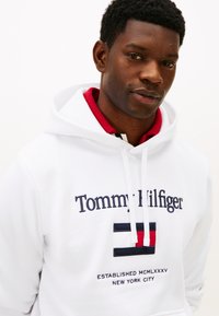 Valkoinen huppari, jossa on kirjailtu "Tommy Hilfiger" -teksti ja logo. Mallissa on kengurutasku ja kiristysnauhalla varustettu huppu. Näkyvillä punaiset ja laivastonsiniset yksityiskohdat.