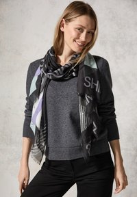 Sciarpa nera e multicolore appoggiata in modo rilassato su un maglione grigio, caratterizzata da motivi geometrici e bordi frangiati. Texture del tessuto morbida.