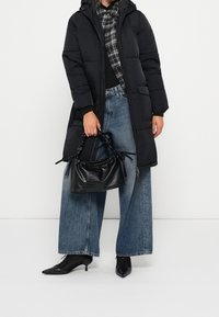 Schwarze Steppjacke mit Kapuze, getragen über einem karierten Hemd, kombiniert mit weit geschnittenen Jeans und schwarzen, schuhlosen Stiefeletten, während sie eine schwarze Handtasche hält.