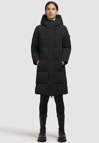 Melna pūkaina mēteļa ar kapuci, taisna piegriezuma, ar izšūtu tekstūru un ar rāvējslēdzēja kabatām. Valkāta ar melnām legingām un kaujas zābakiem.