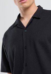Black short-sleeve button-up shirt with a textured pattern, a notched collar, and three black buttons down the front.