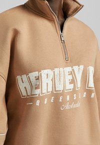 Sweat-shirt brun avec un col montant et une demi-fermeture éclair, présentant un texte blanc qui indique "HERVEY BAY - QUEENSLAND Australie". Texture douce.