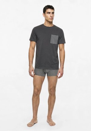 Homme debout pieds nus, portant une chemise gris foncé à manches courtes avec une poche poitrine à motif et un boxer assorti à motif.