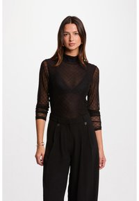 Vrouw met lang bruin haar, gekleed in een zwarte transparante mesh top en hoge zwarte broek, staand voor een vlakke witte achtergrond.