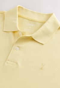 Lysegul poloshirt i tekstureret stof med klassisk krave, tre knapper og et lille broderet logo på brystet.