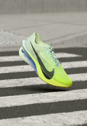 Neon gul og grøn løbesko med sort swoosh, der flyver over et fodgængerfelt på en bygade.