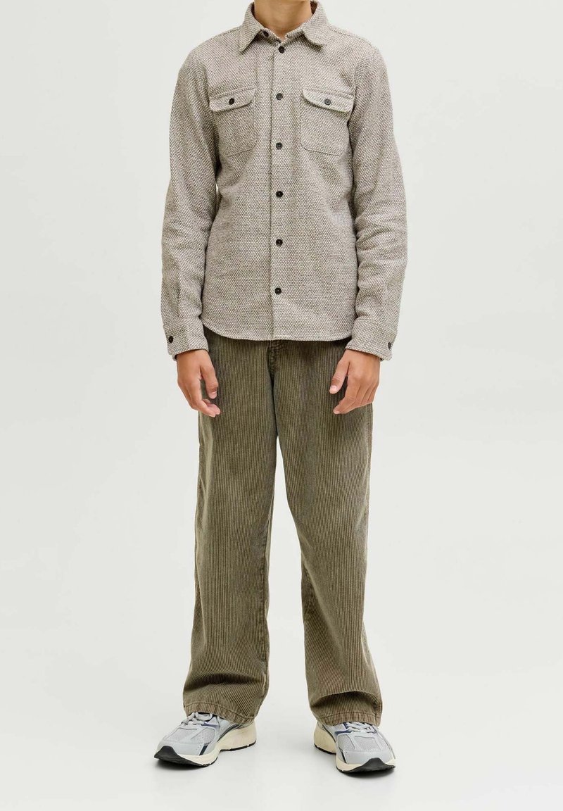 Enfant portant une chemise beige texturée à boutons, un pantalon en velours côtelé vert et des baskets blanches, debout devant un fond clair uni.