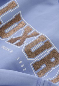 Un tissu bleu clair présentant de grandes lettres en paillettes marron brillantes formant le mot "TUXOR" et une année blanche "1982", avec une texture lisse et douce.