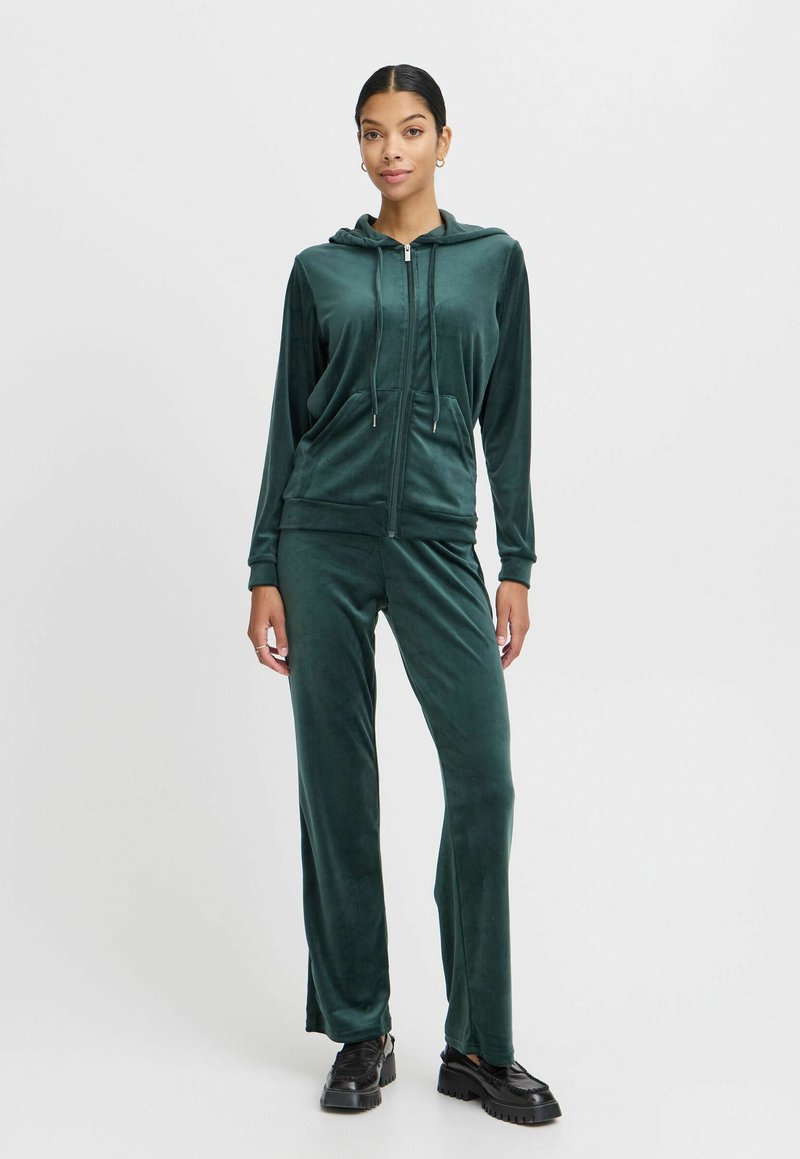 Trening din catifea de culoare verde închis, cu hanorac cu fermoar, buzunare frontale și pantaloni lungi, drepti. Complementează cu încălțăminte cu platformă neagră.