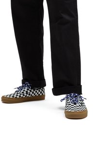 Black pants with cuffed hems paired with low-top shoes featuring a black and white checkerboard pattern and blue laces on a tan sole.